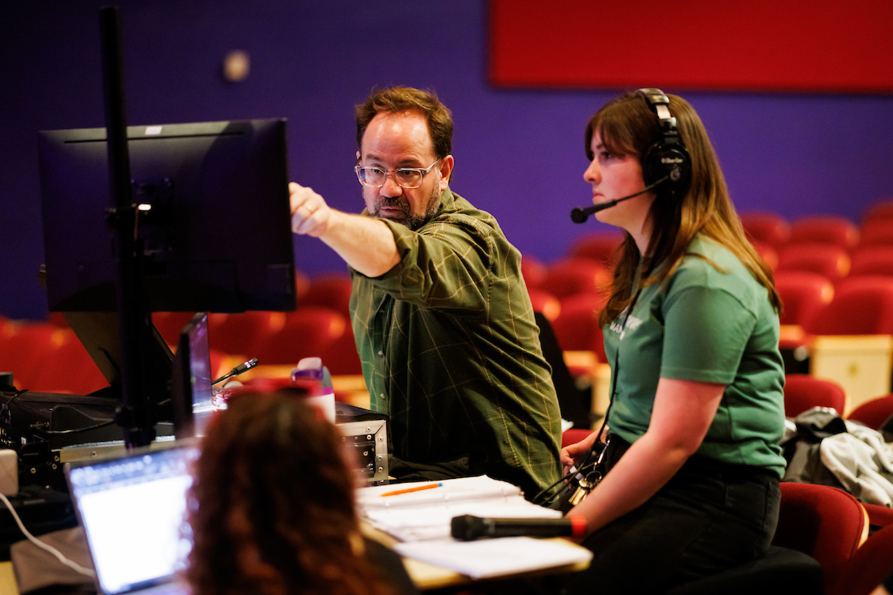 An older man sits next to a young woman wearing a headset, pointing at a monitor at a workstation surrounded by theater seats.