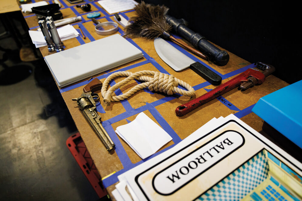 A collection of stage props, including a featherduster, a plastic knife, a wrench, a knotted rope, a revolver, and a large card reading "Ballroom" lie on a table in areas marked off with blue tape.