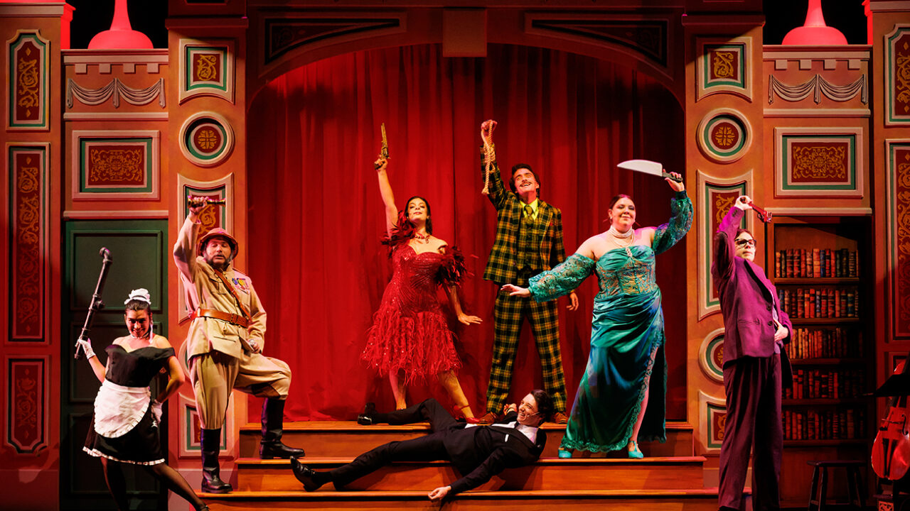 A group of colorfully-dressed characters stand on an elaborate stage set piece, holding up weapons as they surround a young man lying on teh ground wearing a tuxedo.