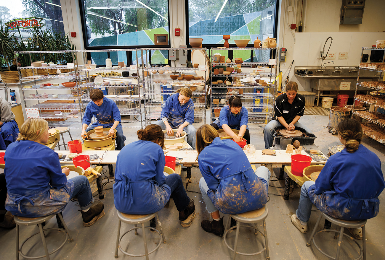 A group of students in blue smocks sit at spinning wheels making pottery.