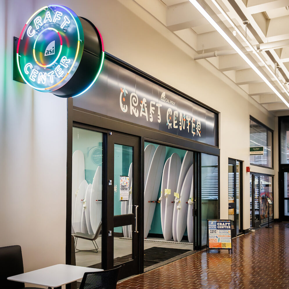 An open glass door in a brightly-lit hallway under colorful signs reading "Craft Center" reveals a rack of surfboards.