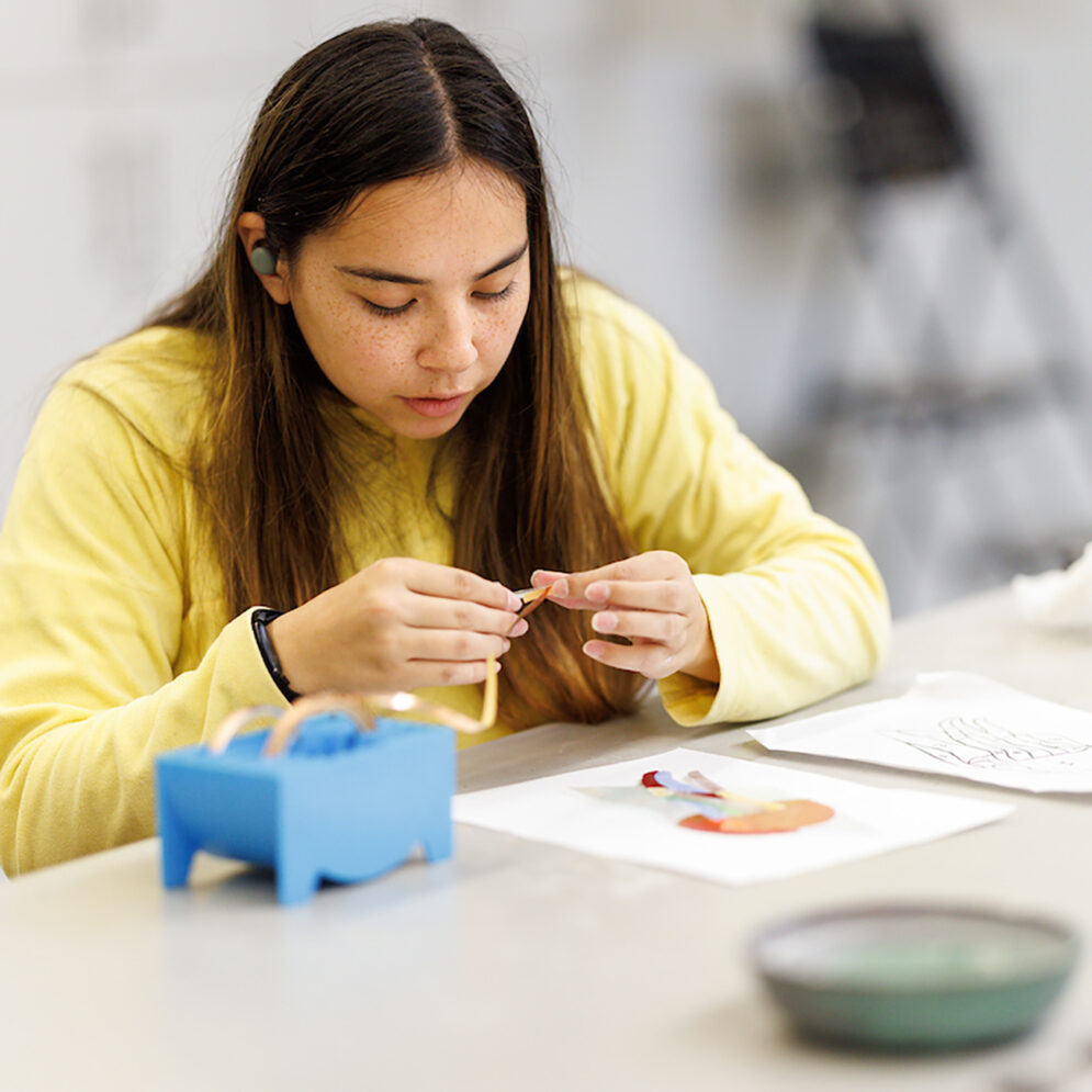 A young woman sits at a table applying a length of metal filament to a small piece of colored glass.