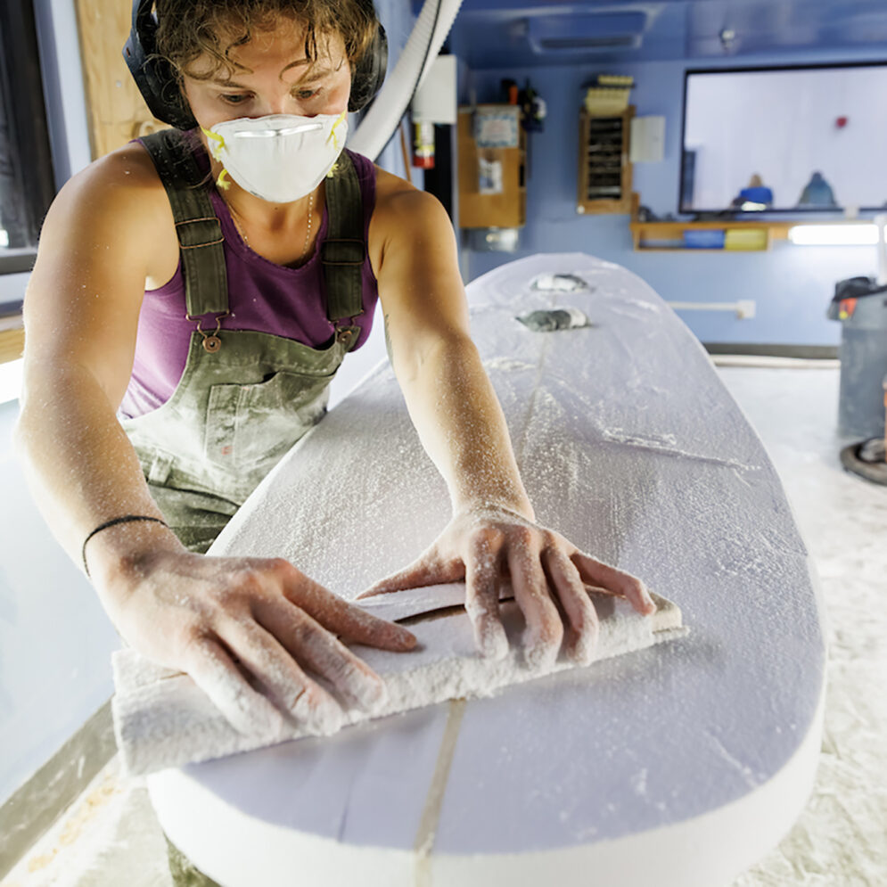 A young woman wearing overalls, a respirator mask and headphones scrapes a tool across the surface of a partially finished foam surfboard blank.