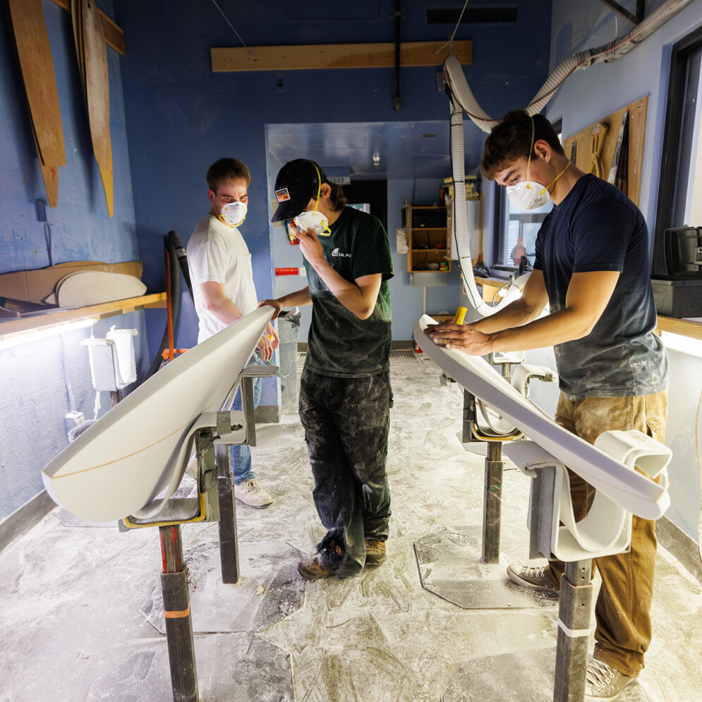 In a workshop strewn with white dust, three young men wearing respirator masks work on shaping foam into surfboards.