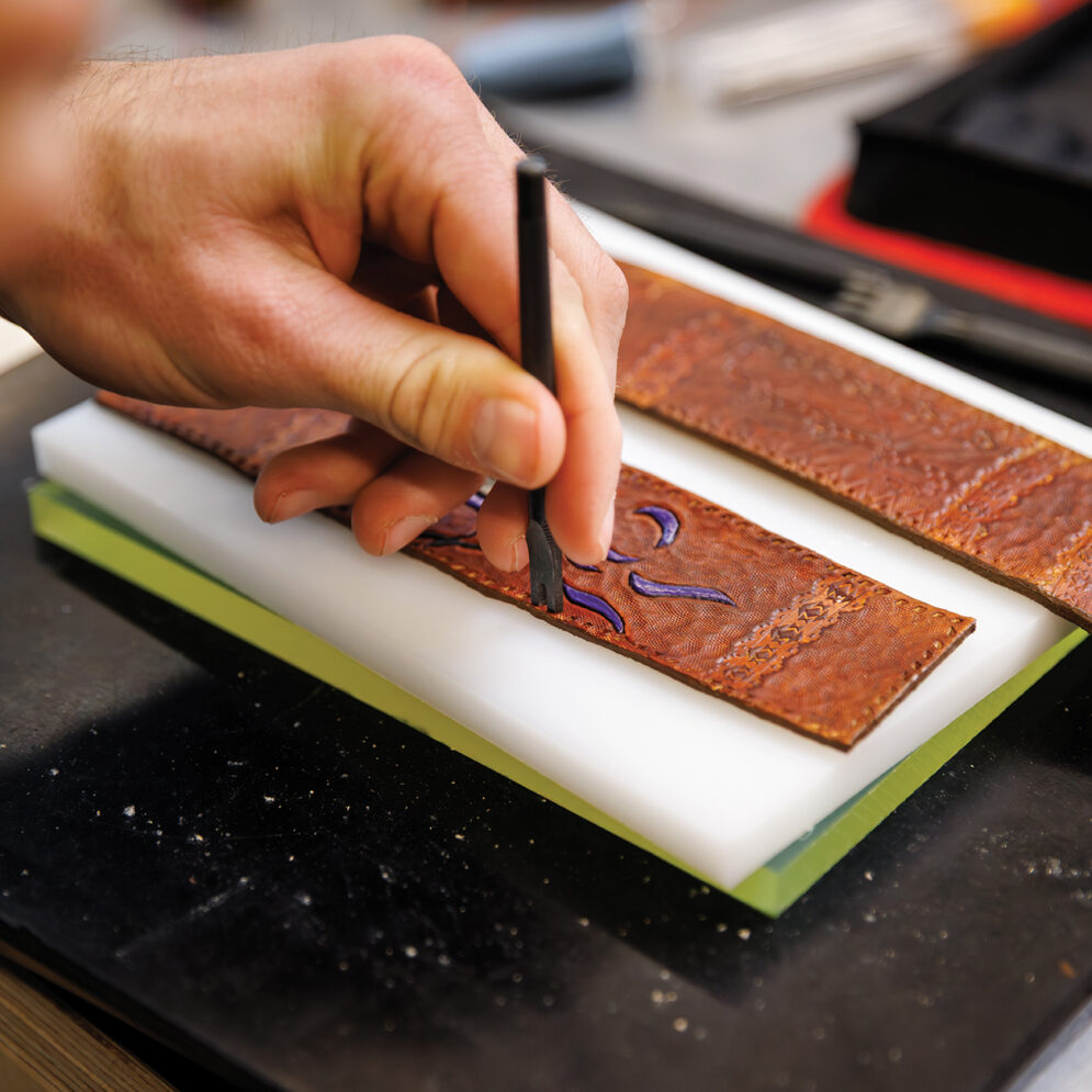 A close-up shot of a hand pressing a metal stamp tool into a decorated leather strap.