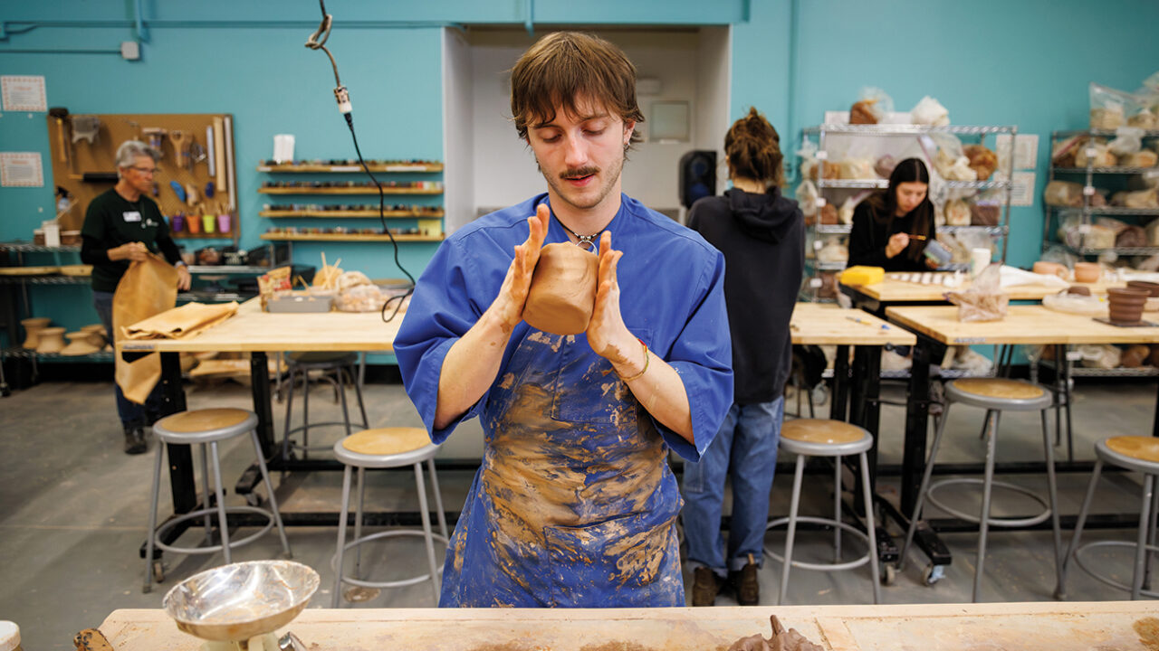 A young man in a blue smock manually shapes a clay pot.