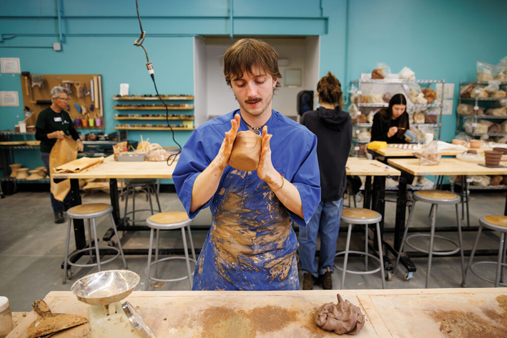 A young man in a clay-spattered blue smock uses his hands to shape a clay pot