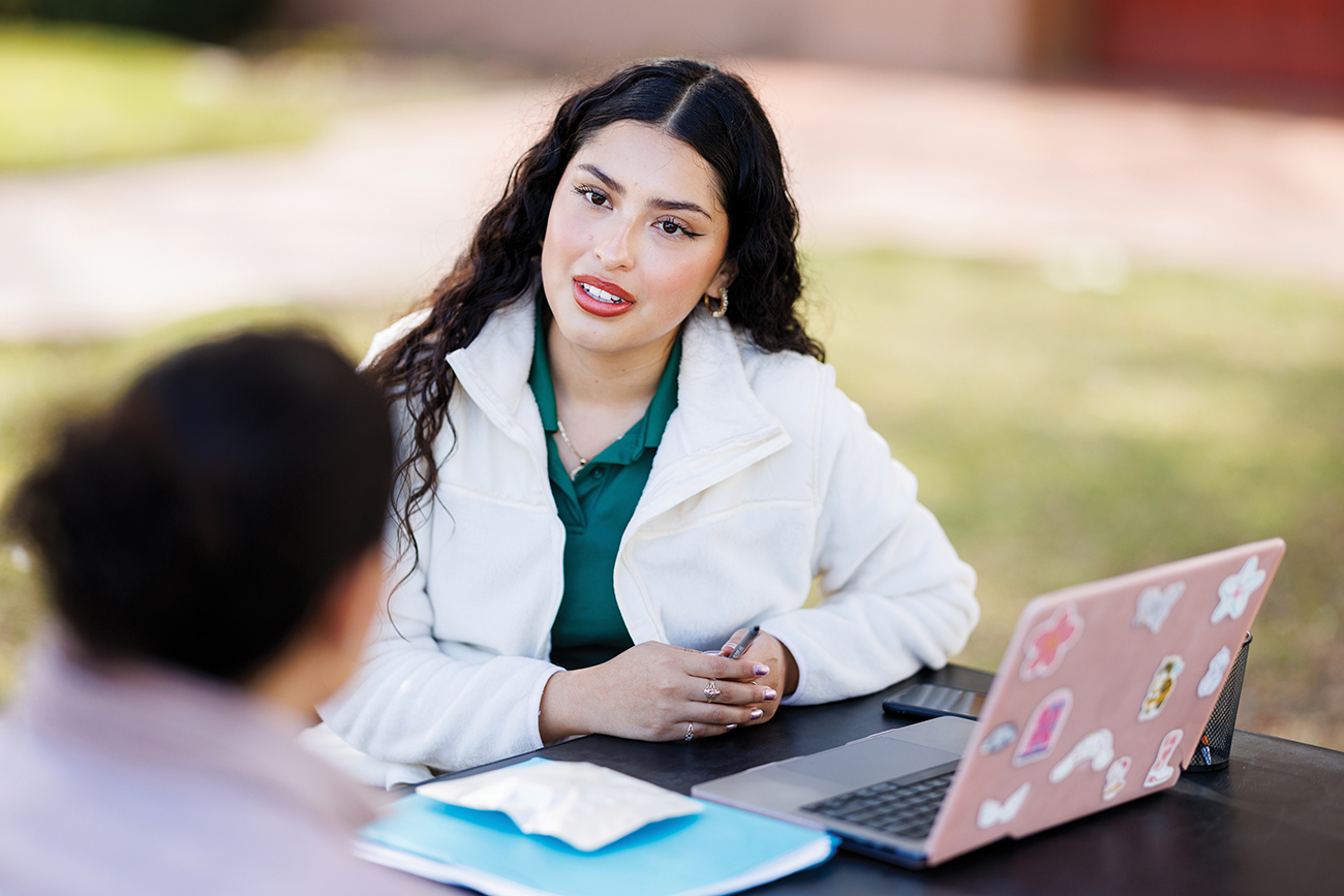 A young woman in a white coat sits in front of an outdoor table with a pink laptop while speaking with another woman.