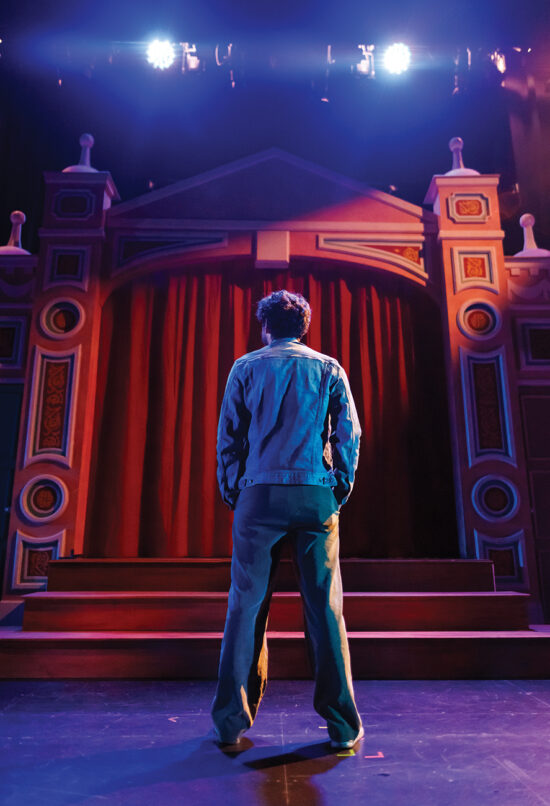 A man in a denim coat stands on a stage looking at an elaborate set piece with spotlights overhead.