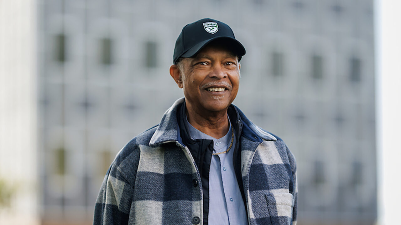 An older man in a cap and plaid shirt stands smiling in front of a tall building.