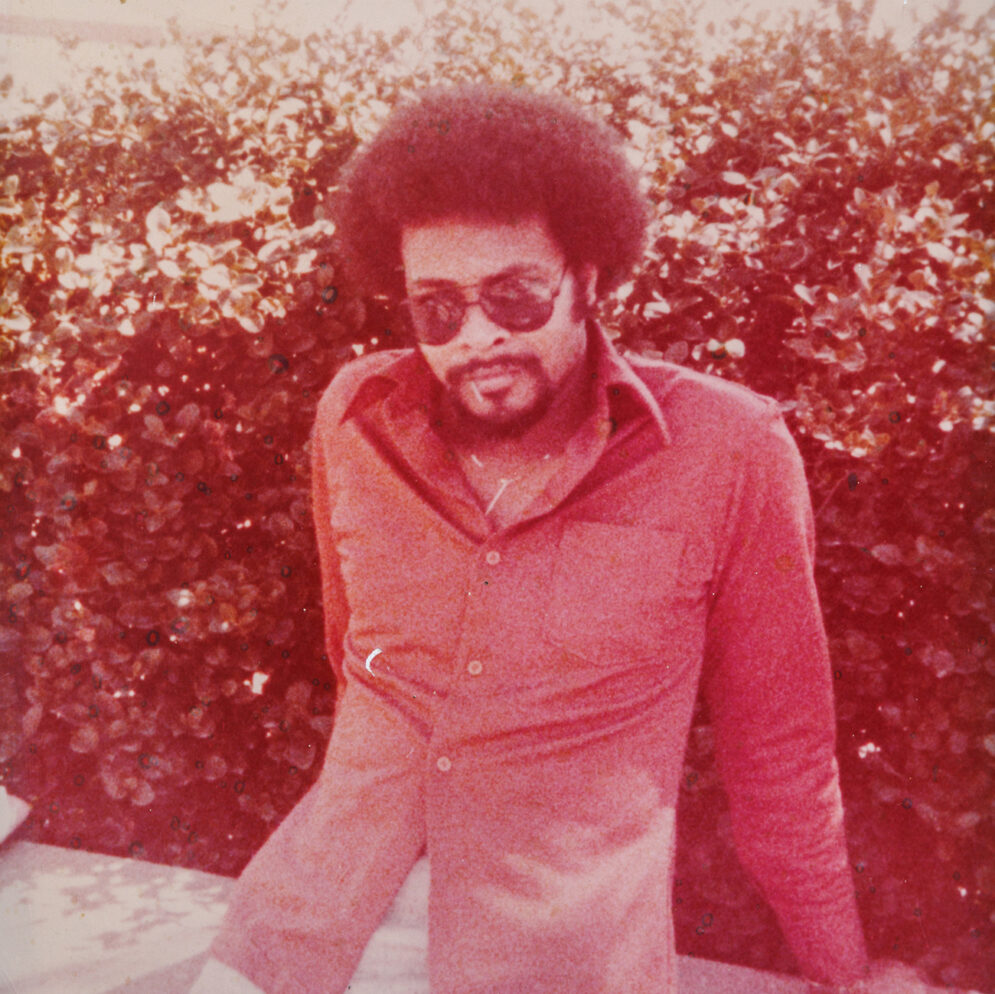 In a vintage photo, a man in a red shirt and sunglasses grins into the camera.