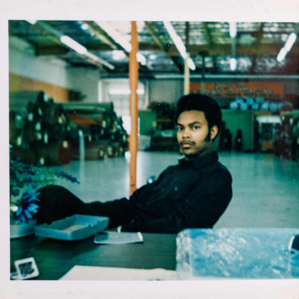 In a vintage photo, a young man in a black coat sits in a chair in a warehouse.