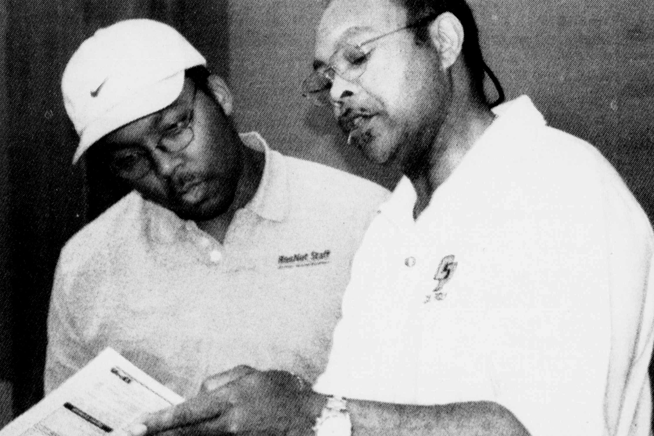 In a vintage black and white photos, a young man in a cap and an older man in a white polo stand together looking at a document.