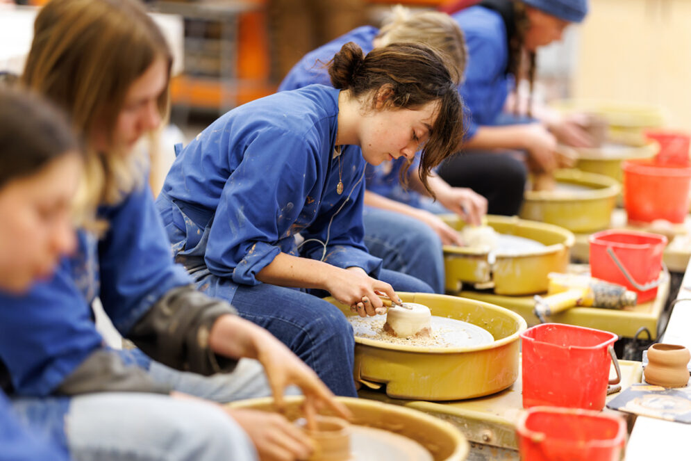 A group of students in dirty blue smocks sit at a row of pottery wheels, shaping pieces of clay.