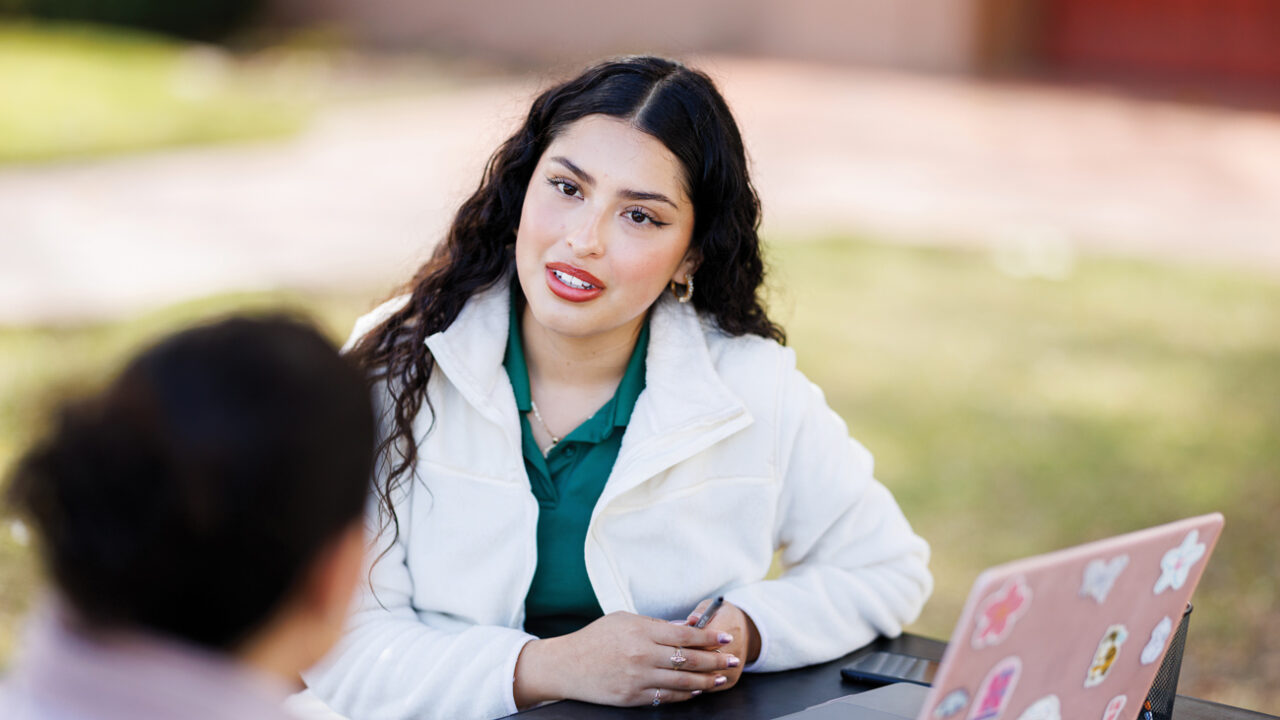 A student in a white lab coat with a laptop talking to a patient