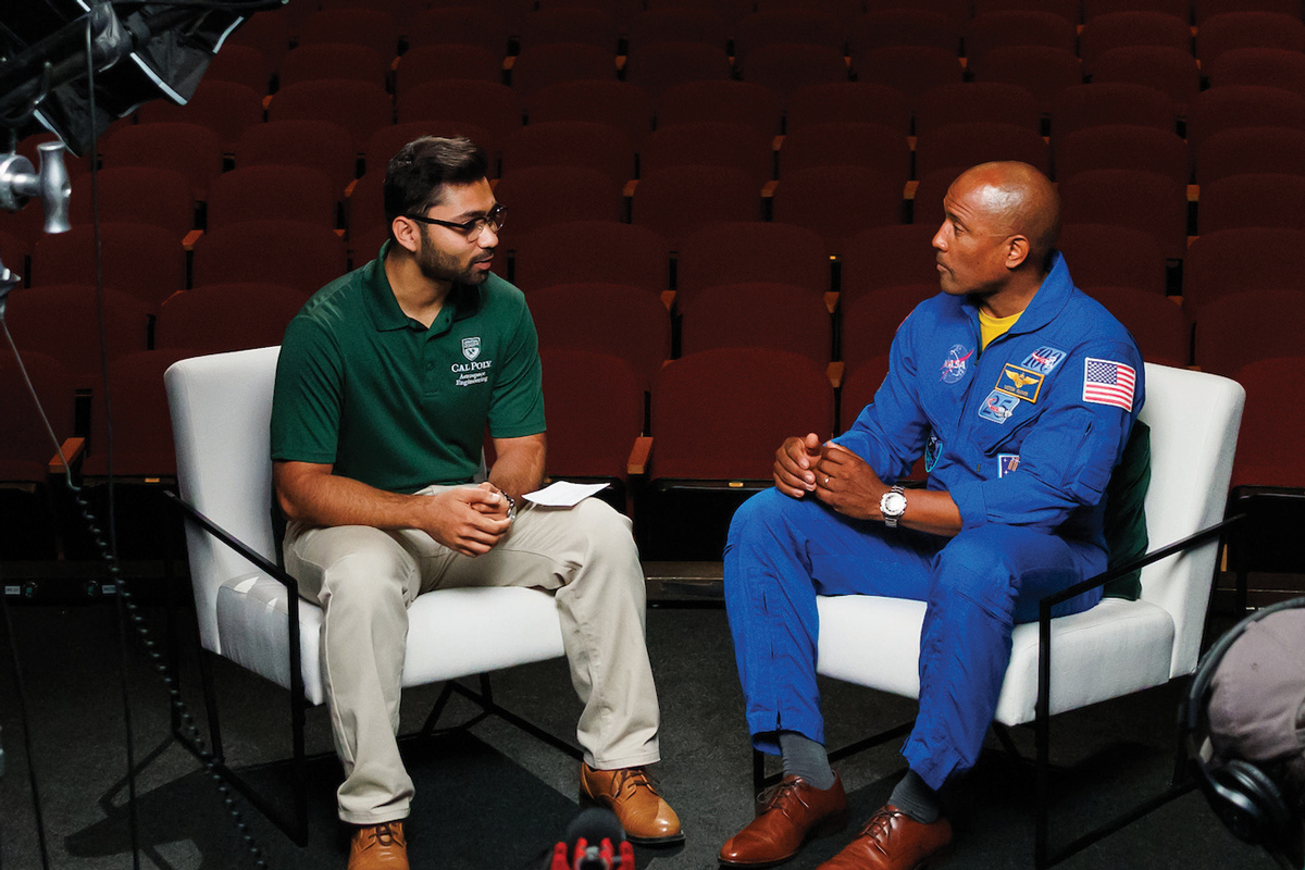 A Cal Poly student and NASA astronaut Victor Glover sit in white chairs on the stage of a theater on a video shoot set