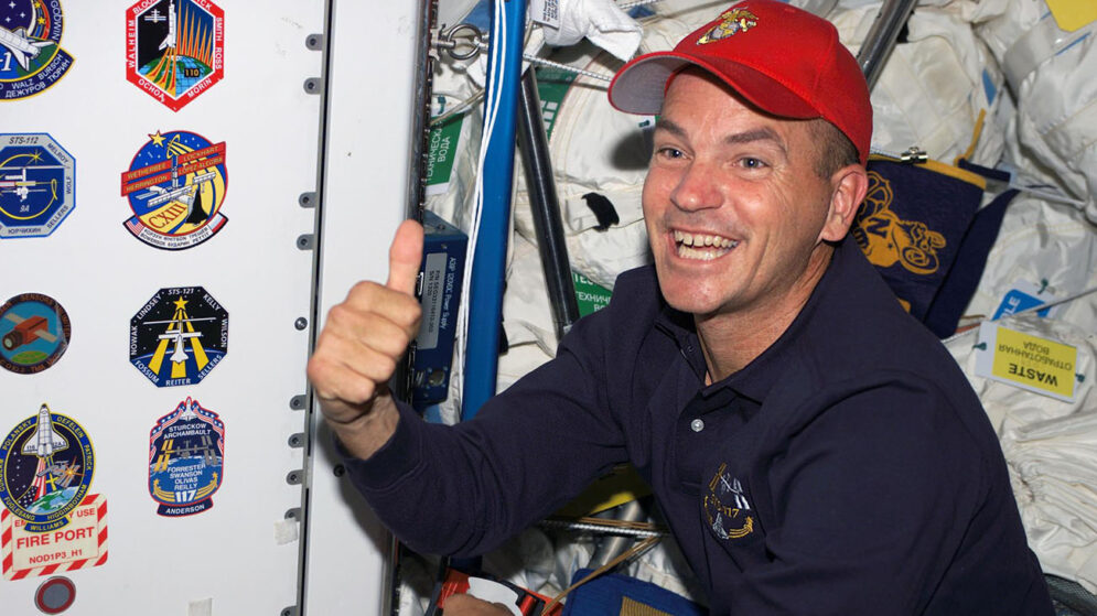 A man in a blue polo shirt and red cap gives a thumbs-up gesture as he floats in front of a wall displaying NASA space mission insignia