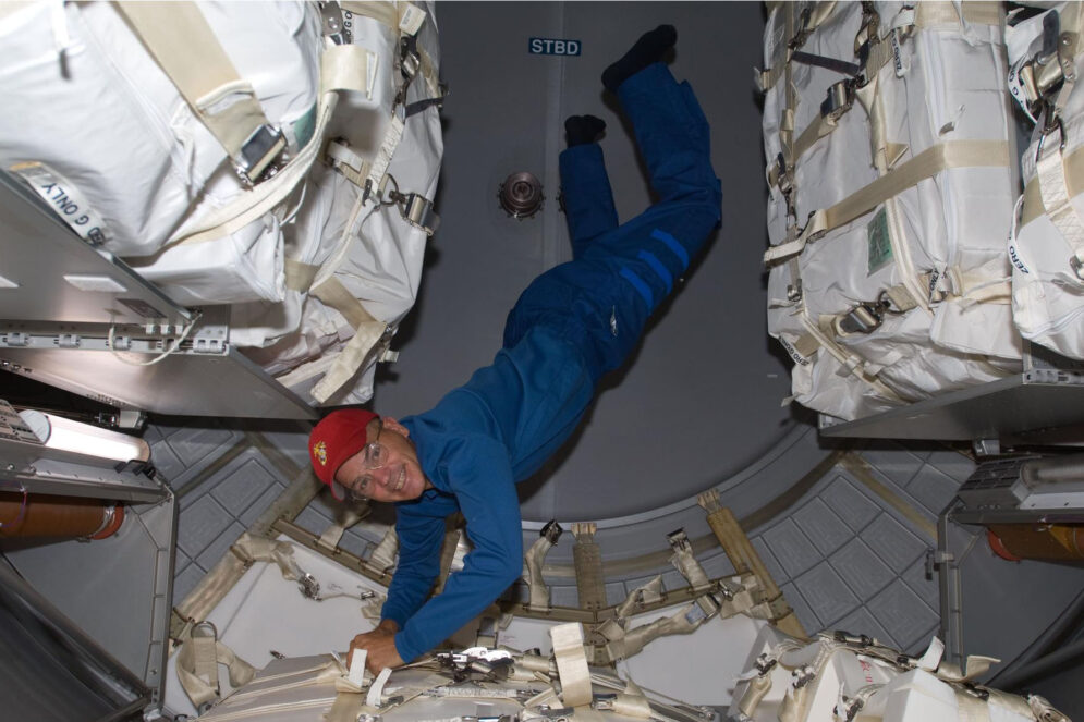 A man in a blue jumpsuit and red cap floats upside down in a spacecraft laboratory