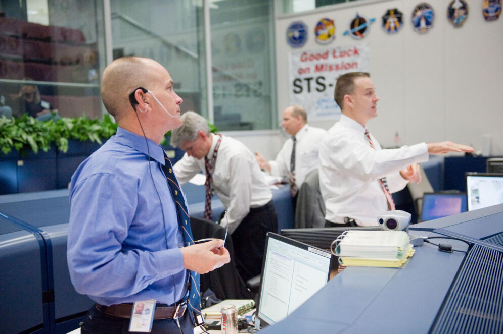 A man in a blue shirt and tie wears a communications headset in a room full of computer monitors and NASA insignia