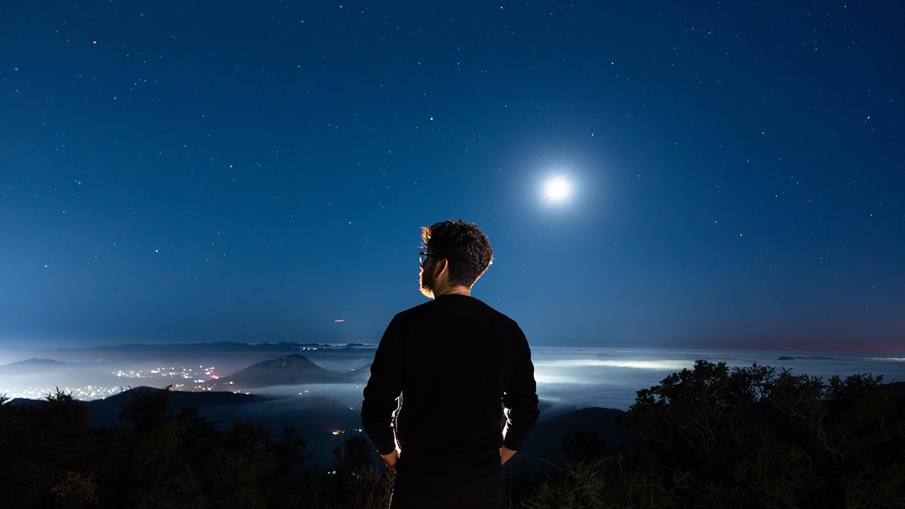 A man stands on a hilltop looking into a night sky filled with stars and a full moon.