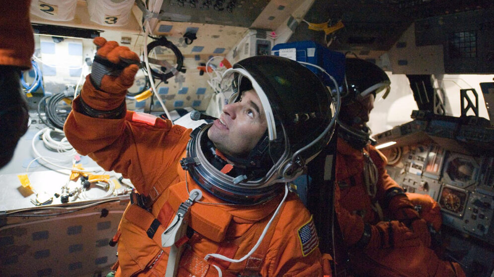 A man in an orange suit and space helmet looks up at mounted equipment in a mock spacecraft