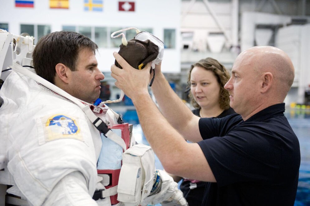 A man in a polo shirt helps put a headset on another man in a bulky space suit