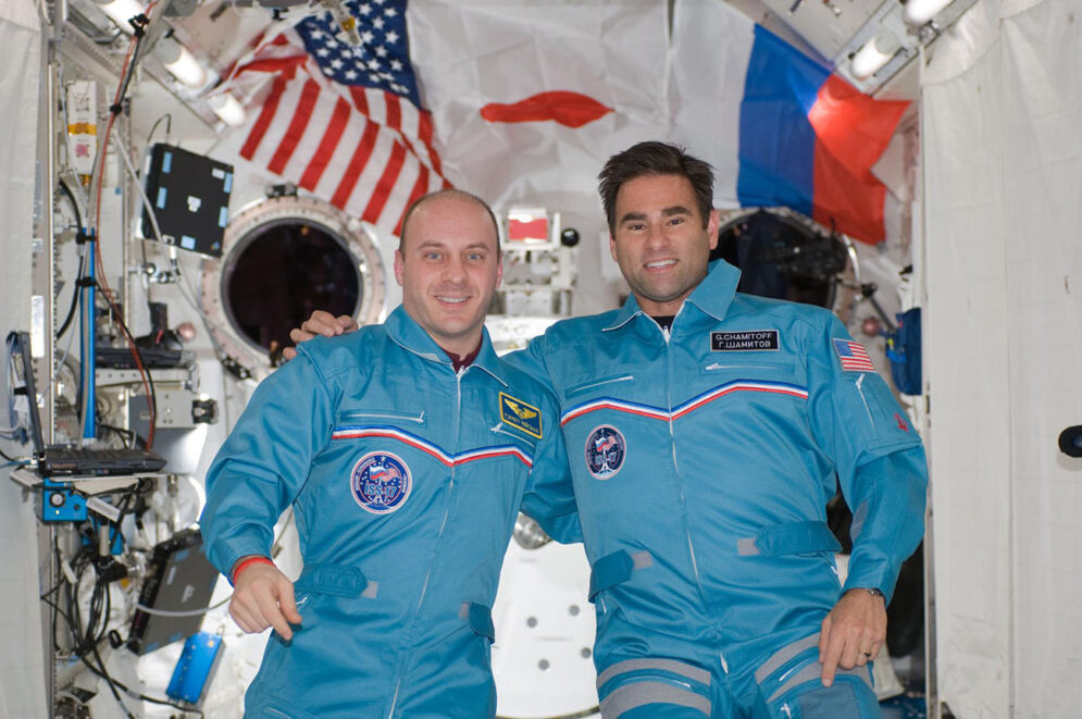 Two men in blue jumpsuits smile for a photo as they float together in a space station laboratory with U.S., Japanese and Russian flags displayed in the background