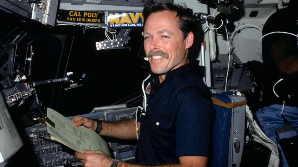 A smiling man sits among the control panels in a spacecraft, with stickers reading "Cal Poly San Luis Obispo" and "Navy" pasted on the wall behind him.