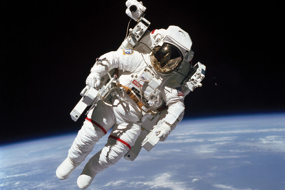 An astronaut in a white space suit and helmet floats freely in space with the curved surface of the Earth in the background