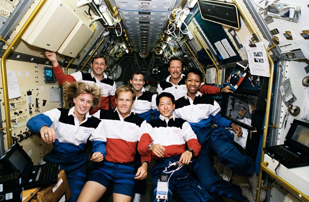 A group of men and women smile for a portrait as they float together through the air inside a spacecraft