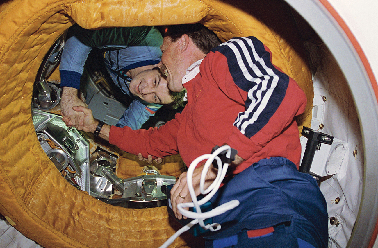 Two men floating in midair smile as they shake hands through the portal of a spacecraft