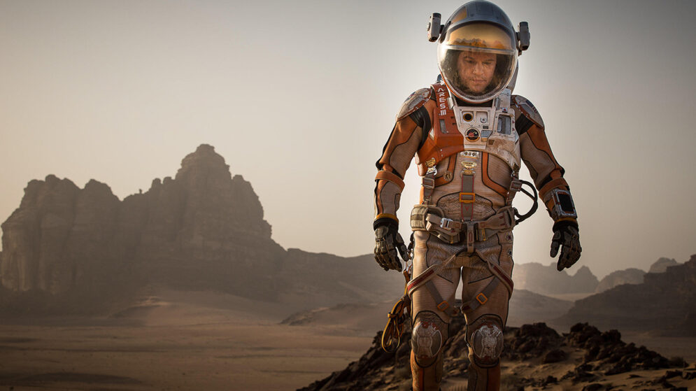 A man in an orange and white space suit and helmet walks through a desert landscape.