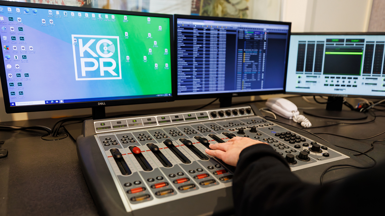 A hand on a sound control board in front of a bank of monitors displaying music playlists and the KCPR logo