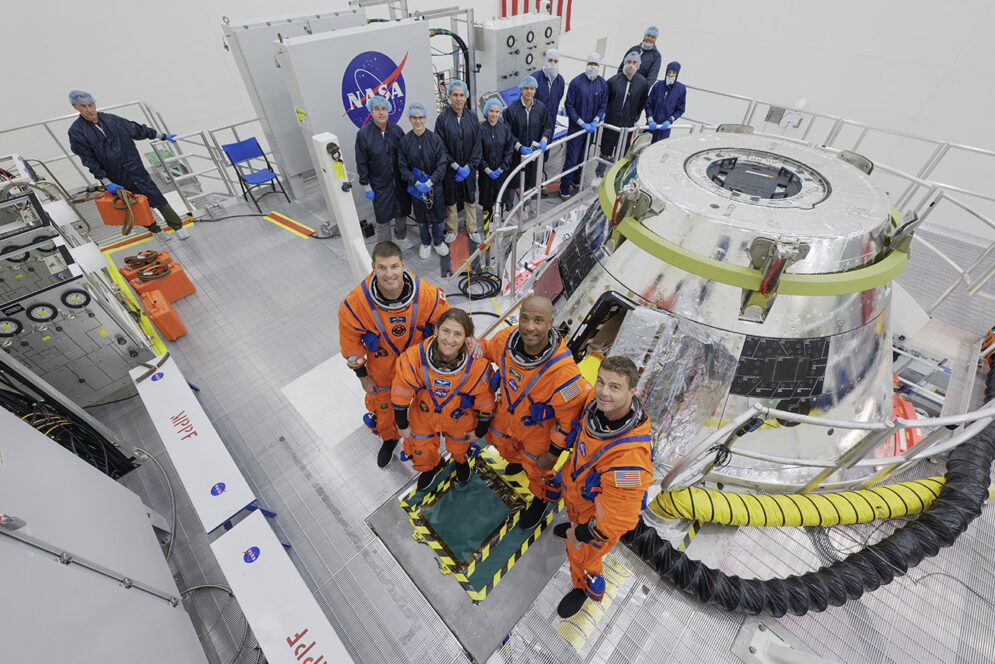 Four people in orange jumpsuits pose for a photo in front of a space capsule with a group of technicians in blue coveralls in the backgrounf