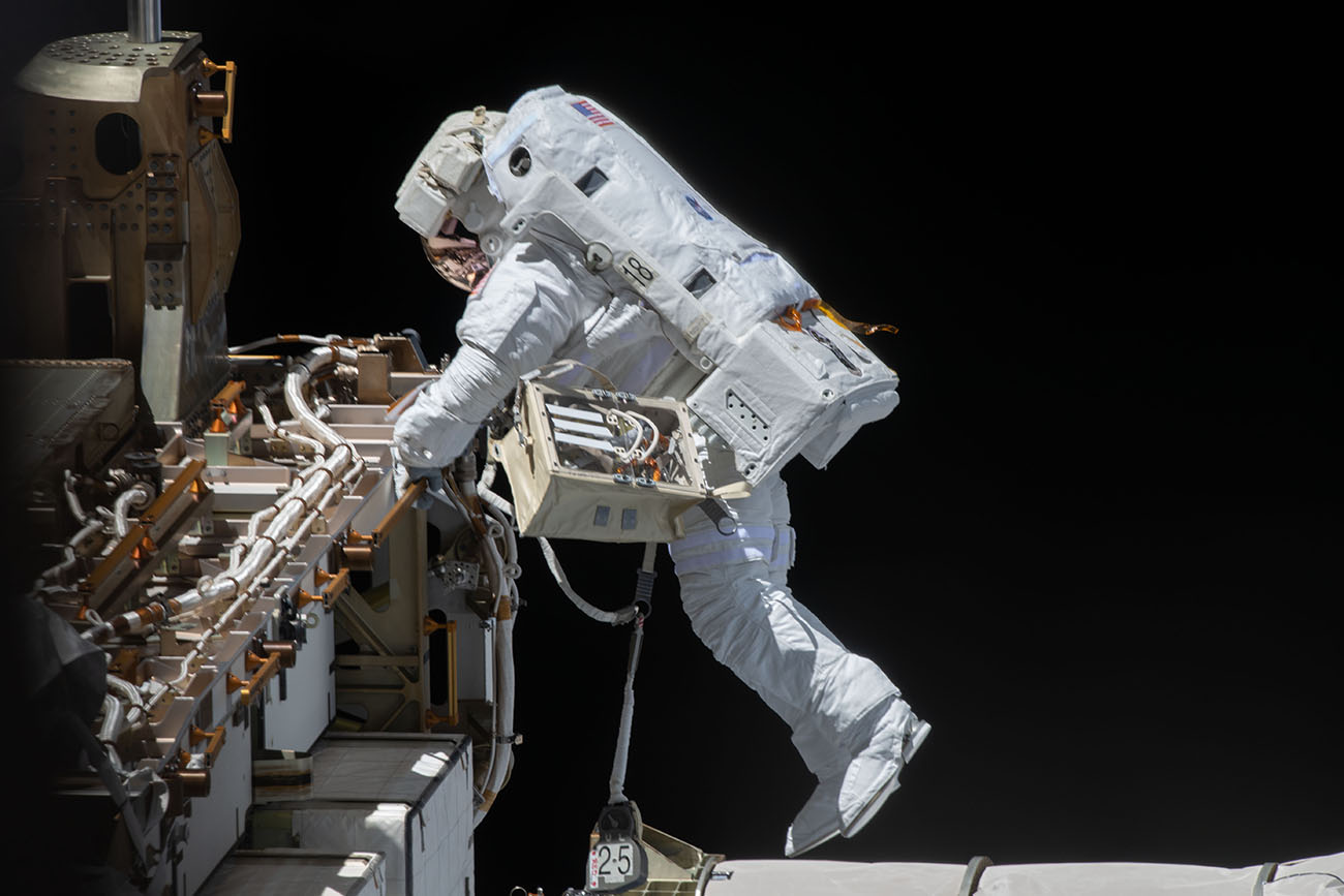 An astronaut in a white suit and helmet makes a repair to the exterior of a space station