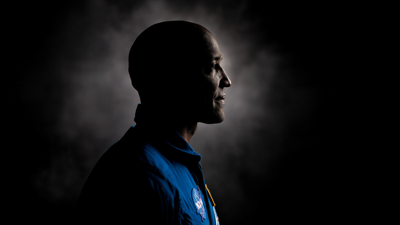 A profile portrait of a man in a blue jumpsuit with NASA patches against a dark background