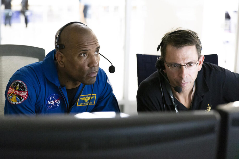 Two men wearing communications headsets monitor a bank of computer stations
