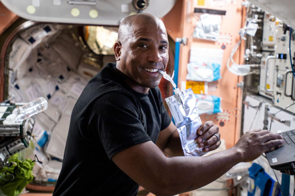 A man in a black T-shirt sips from a foil pouch as works on a laptop while floating in a space station