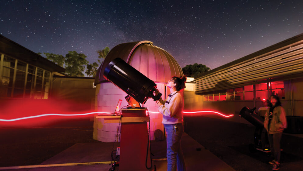 A student looks up at a starry night sky in front of a telescope and a small, domed observatory