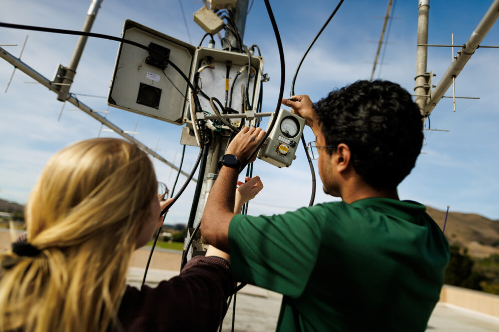 Two students hold wires and sensors near an antenna on the roof of a building