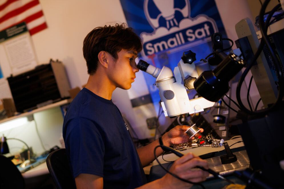 A student looks through a microscope while soldering a circuitboard in a lab that has a Small Sat poster behind him