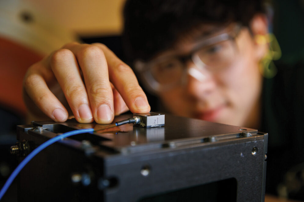 A student wearing glasses places a censor on the surface of a CubeSat payload before testing