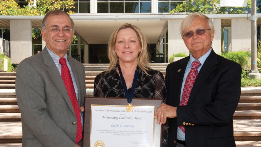 Two people wearing suits present a plaque and medal to a woman