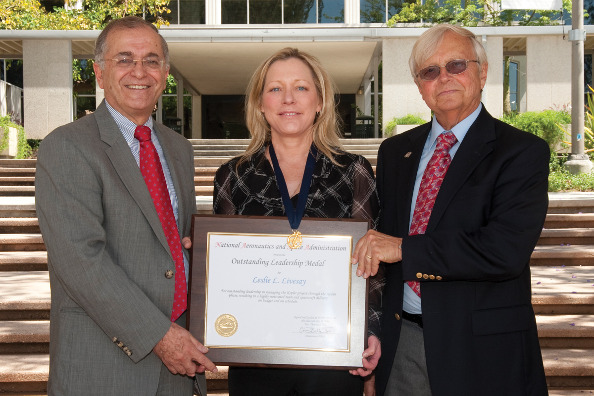 Two people wearing suits present a plaque and medal to a woman