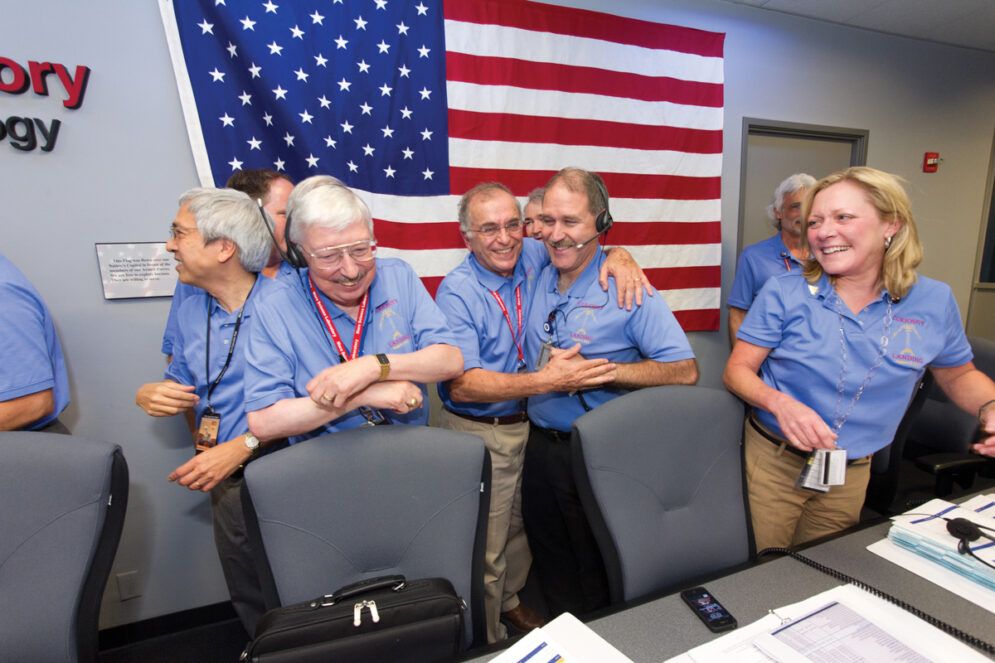 A group of people in blue polo shirts smile and hug in front of an American flag in Jet Propulsion Laboratory's mission control center