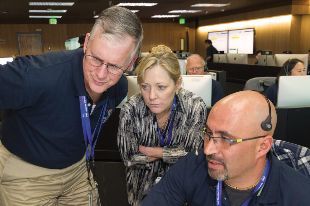 Three people wearing blue lanyards and ID badges look closely at a computer screen.