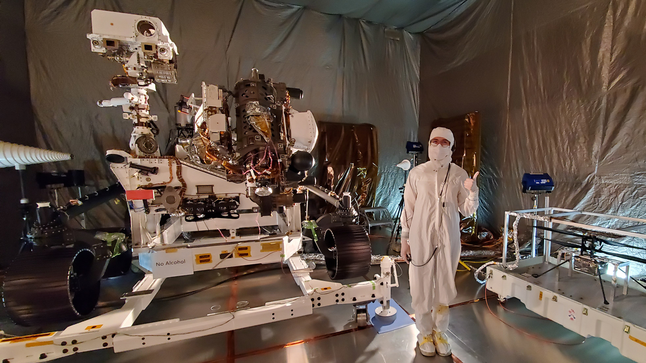 The Mars Perseverance rover in a clean room with an engineer wearing white coveralls and a mask while giving a thumbs up