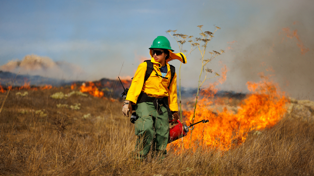A young woman in protective gear holding a drip torch stands in front of a column of flame burning a dry, grassy hillside.