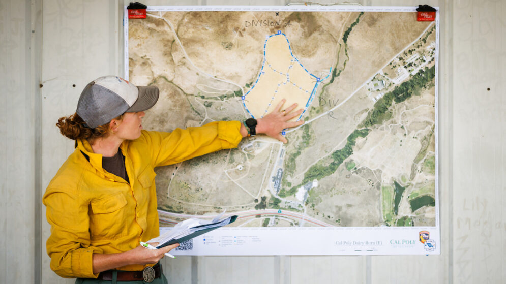 A woman in a yellow jacket and cap gestures to a map mounted on a white wall.