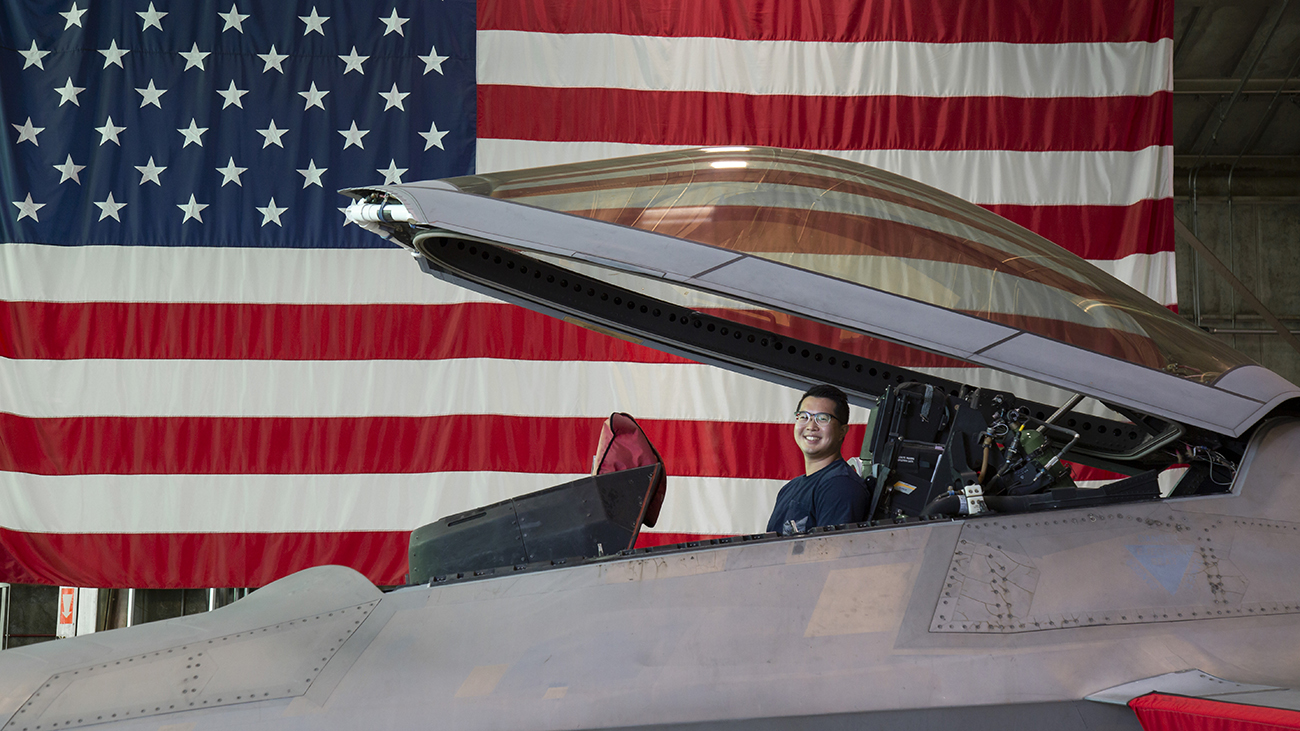A man in a polo shirt smiles in the open cockpit of a fighter jet, with the American flag in the background.